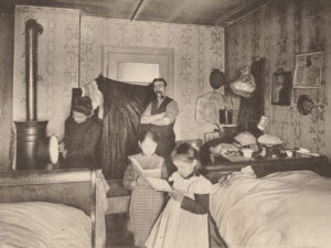 Die beengten Wohnverhältnisse dieser Zürcher Heimarbeiterfamilie bedingen die Nutzung eines einzigen Raums als Küche, Wohn- und Schlafzimmmer. Postkarte anlässlich der Schweizerischen Heimarbeitsausstellung, um 1900.