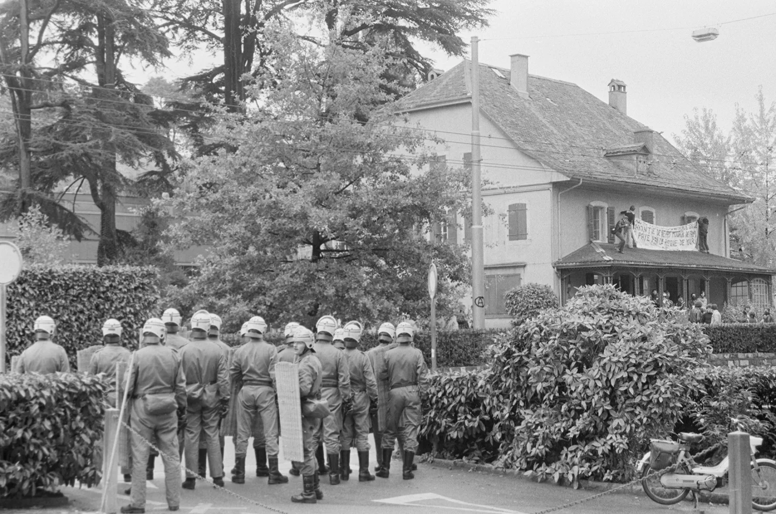 Zwei Personen hängen vom Terrassendach des besetzten Hauses aus das Transparent auf, während die Polizei abwartet. Die dritte Person auf dem Dach filmt beides.