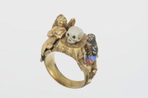 Victorian ring with miniature still-life sculpture of a skull on a table, surrounded by an angel and an owl, as well as butterfly. The ring dates from about 1890 to 1900.