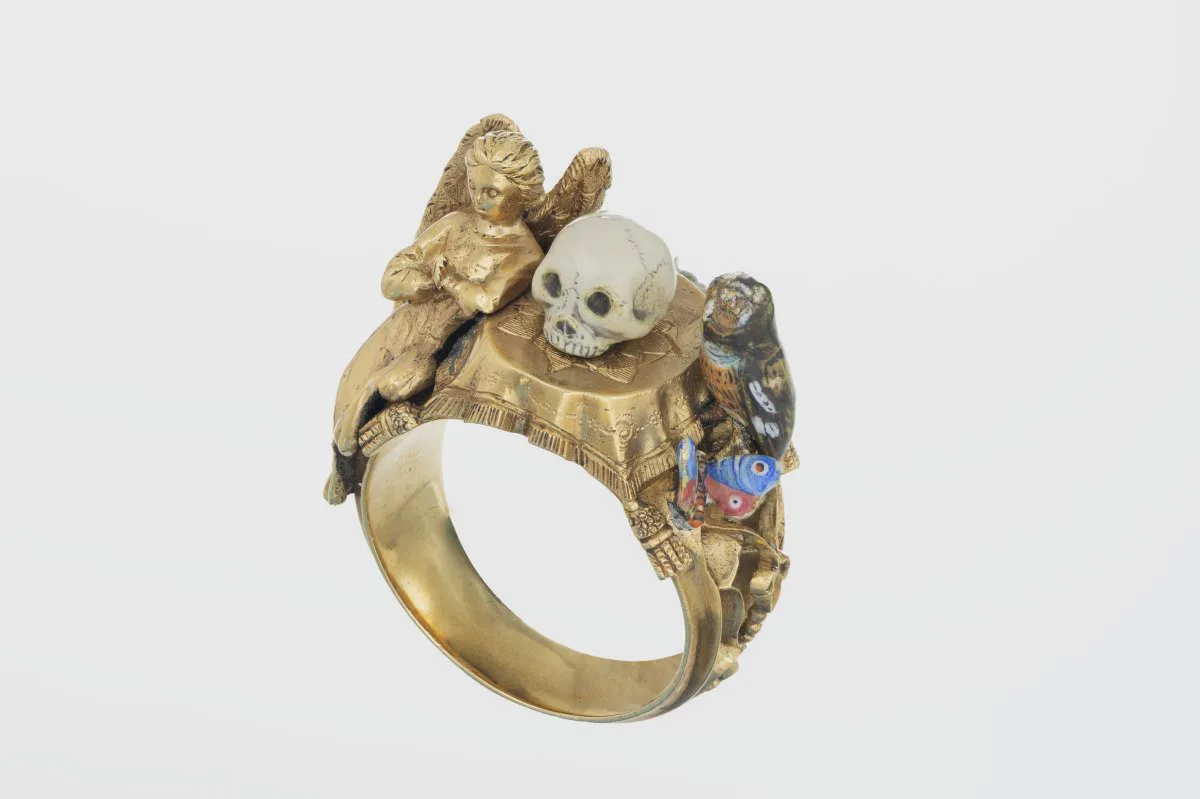 Viktorianischer Ring mit der Miniatur-Stillleben-Skulptur eines Schädels auf einem Tisch, umgeben von einem Engel und einer Eule sowie einem Schmetterling. Der Ring stammt aus der Zeit um 1890 bis 1900.