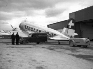 Remorqueur électrique à trois roues Swissair de l’EFAG pour les avions derrière un DC-2, à Dübendorf. À côté, un véhicule de livraison Peugeot, en 1937.