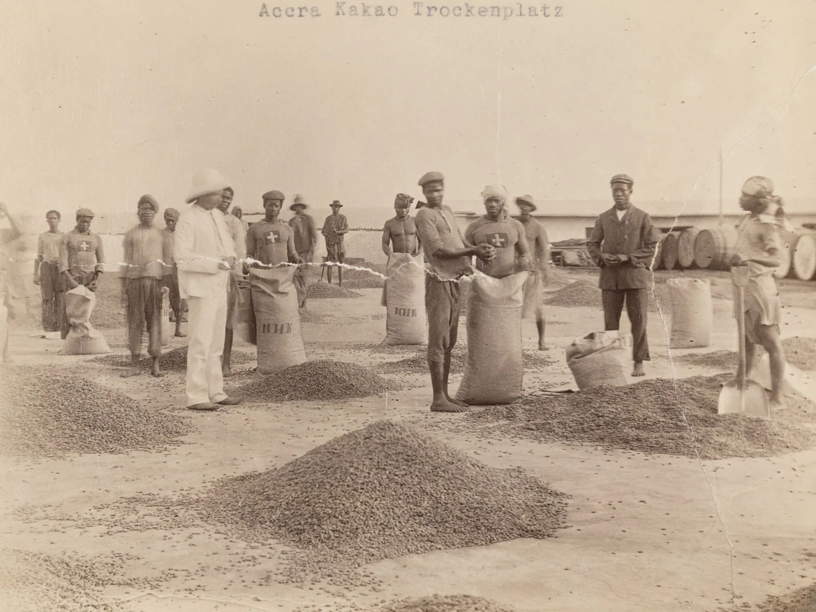 Des travailleurs locaux et un contremaître en tenue coloniale sur une place de séchage des fèves de cacao appartenant à la Basler Missionshandelsgesellschaft, à Accra, dans l’actuel Ghana (1904/1905).