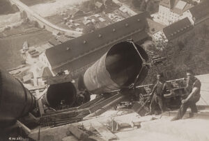 Abladen eines Rohres von der Standseilbahn für die Konstruktion der Druckleitung. Im Tal unten ist das Wasserkraftwerk Amsteg zu sehen. Aufnahme um 1920.