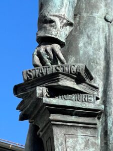 He holds a manuscript in his right hand, while his left rests on three books that embody his achievements: STATISTICA, [STORIA], ISTRUZIONE. Stefano Franscini monument in Faido (detail).