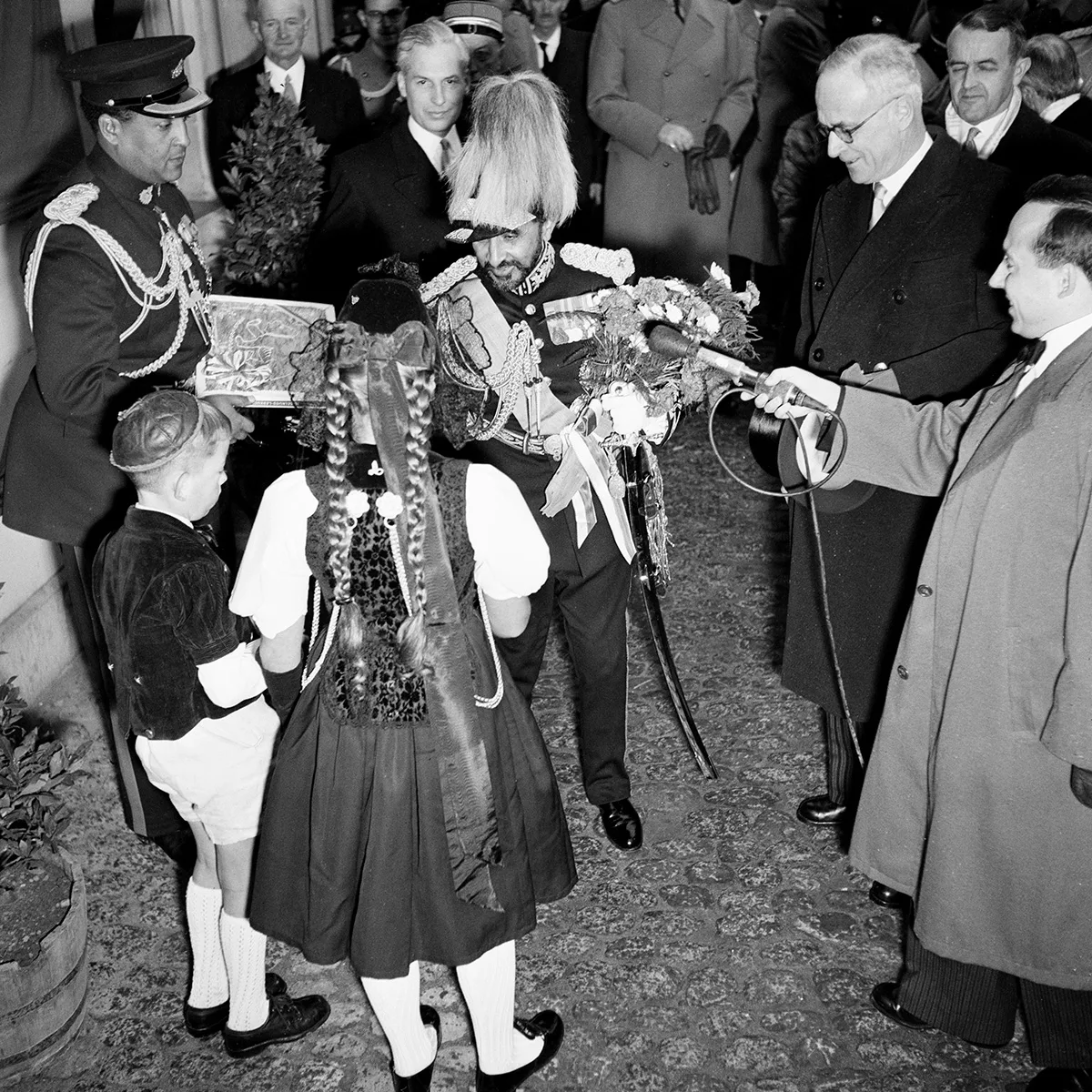 Ein und Trachtenmädchen und ein Bub im Kühermutz überreichen dem Kaiser einen Lebkuchen. Als Gegengeschenk erhalten sie vom Kaiser Münzen.